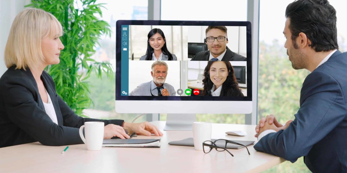 A female and a male manager meet in person while video conferencing their team members.