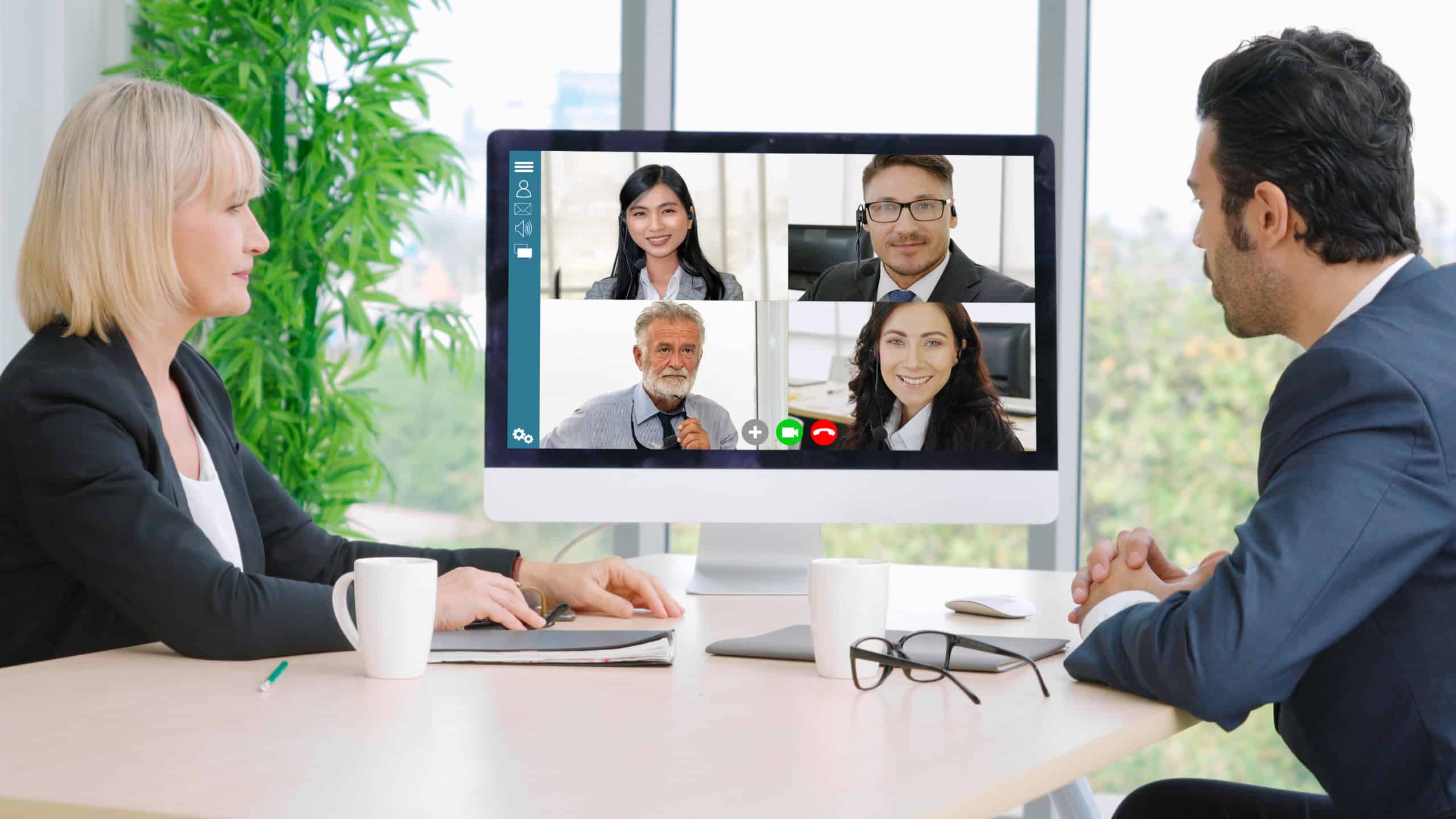 A female and a male manager meet in person while video conferencing their team members.